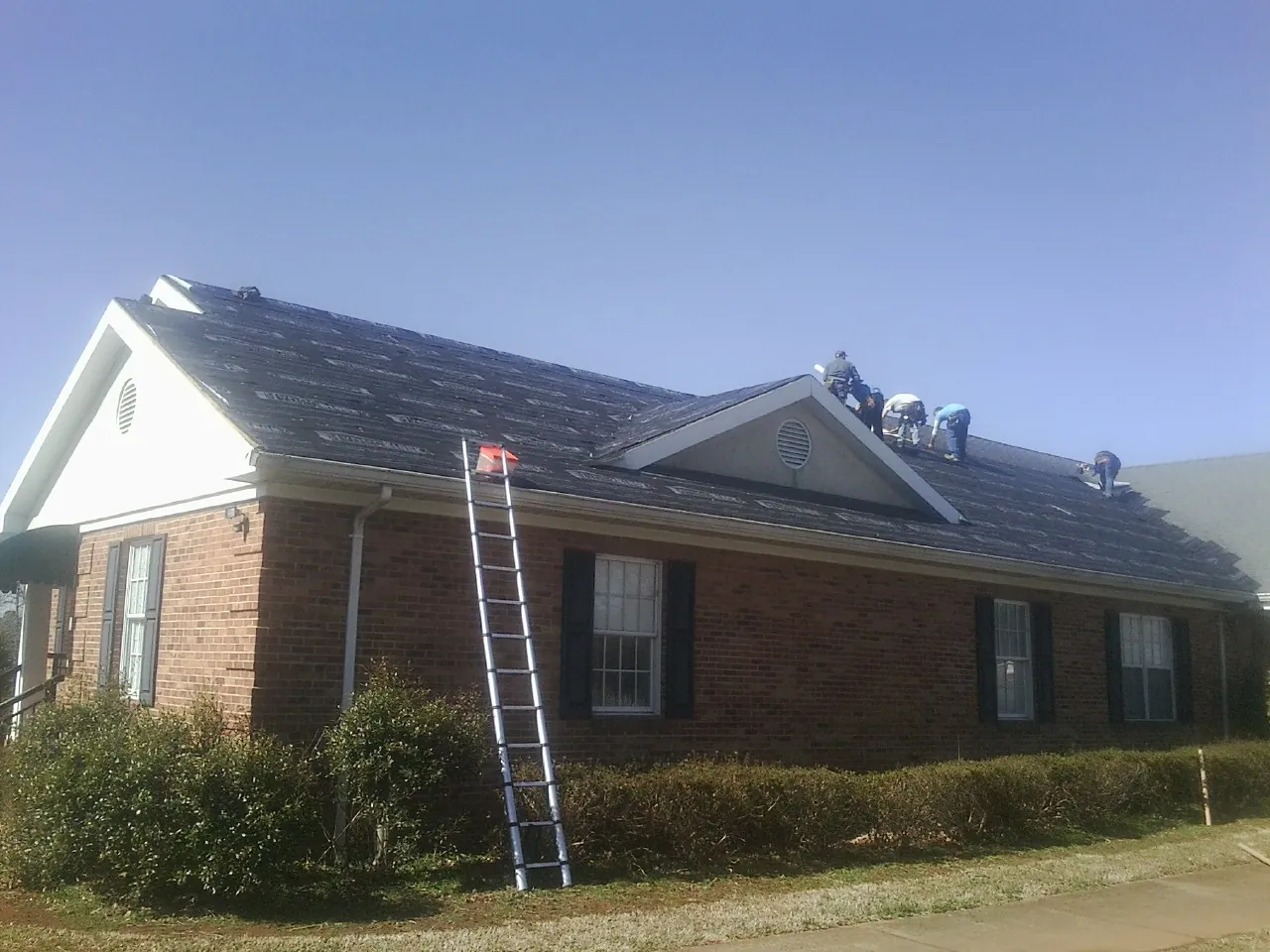 Crew tearing shingles off a brick ranch roof with a ladder set — Metal Roof Repair work by our crew in Lyon Station