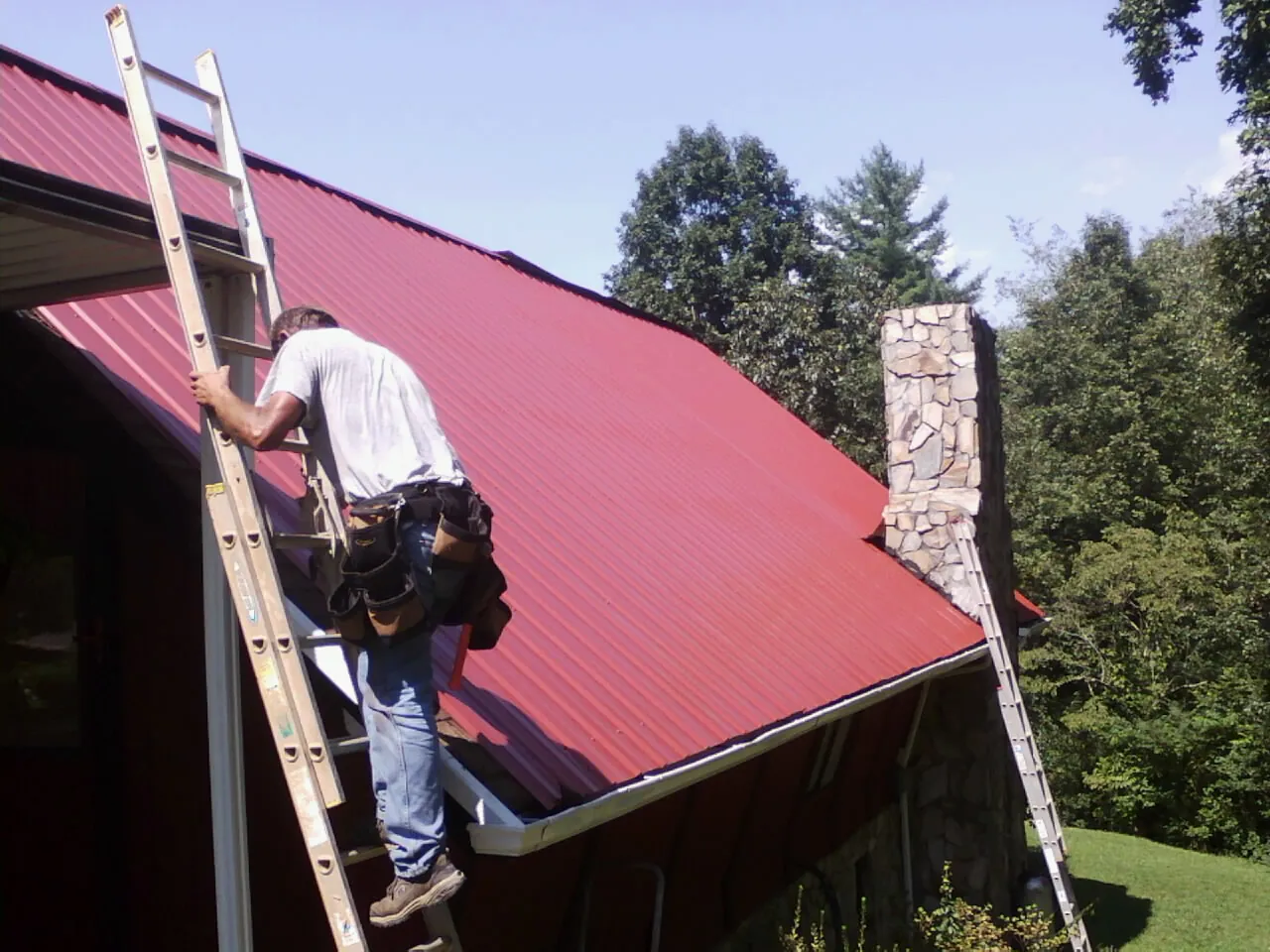 Roofer on a ladder beside a red metal roof and stone chimney — Soffit & Fascia Repair by Roofing Brothers in Lyon Station